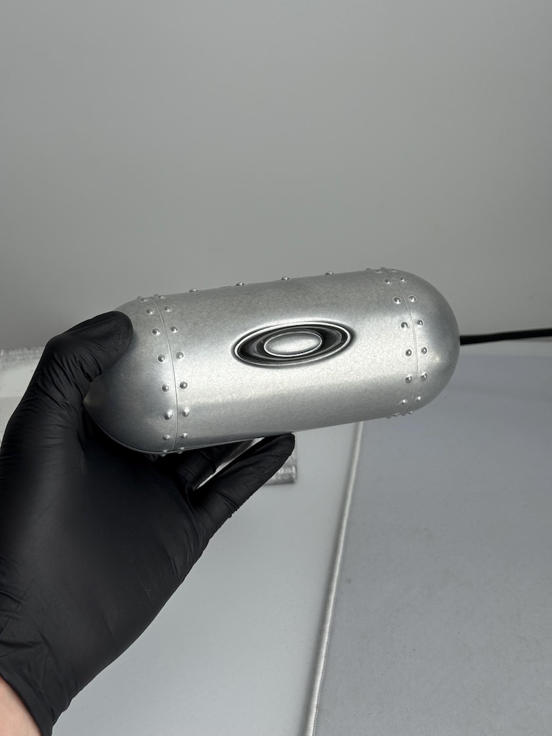 May include: A silver-colored Oakley case with a rounded, cylindrical shape. The case features the Oakley logo and small rivet details. The case is held by a gloved hand.