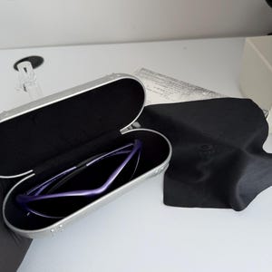 May include: A pair of purple sunglasses rests inside a silver case with a black interior. A black microfiber cloth and a white paper are next to the case. The sunglasses have a sleek, modern design.