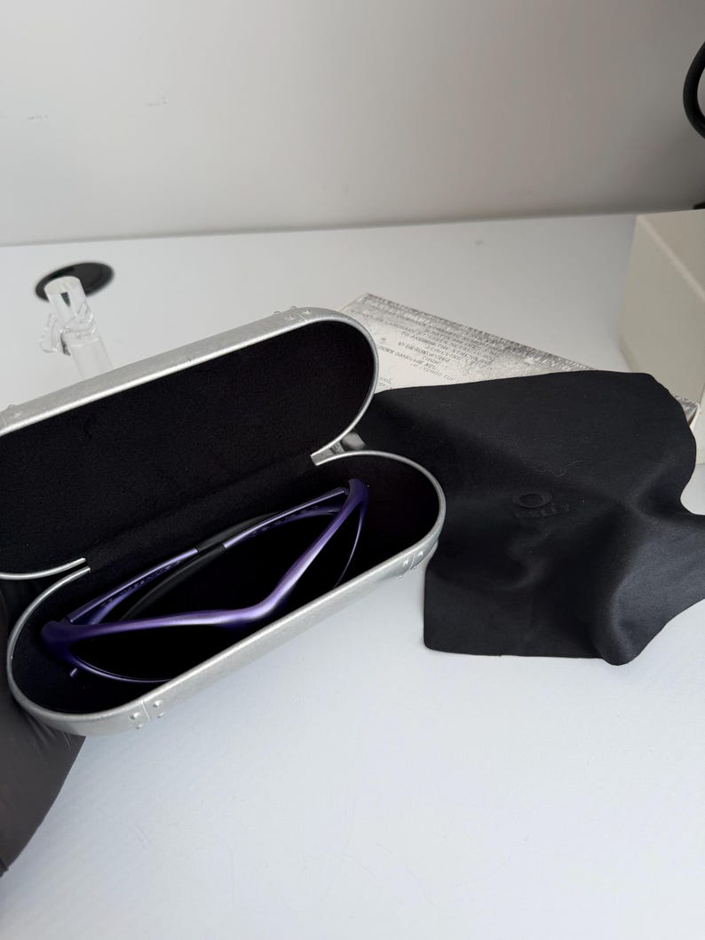 May include: A pair of purple sunglasses rests inside a silver case with a black interior. A black microfiber cloth and a white paper are next to the case. The sunglasses have a sleek, modern design.