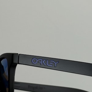 May include: Black sunglasses with blue lenses. The word "OAKLEY" is printed in purple on the temple. The frame is matte black. The sunglasses are designed for outdoor activities.