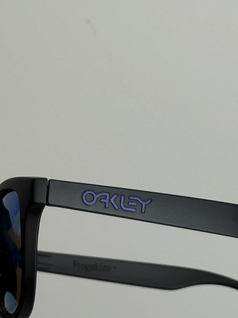 May include: Black sunglasses with blue lenses. The word "OAKLEY" is printed in purple on the temple. The frame is matte black. The sunglasses are designed for outdoor activities.