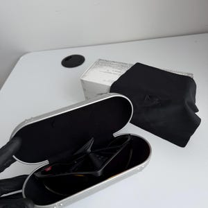 May include: Black sunglasses with a silver case, a black microfiber pouch, and a white box. The sunglasses are in an open silver case with a black interior. The black pouch has the Oakley logo.