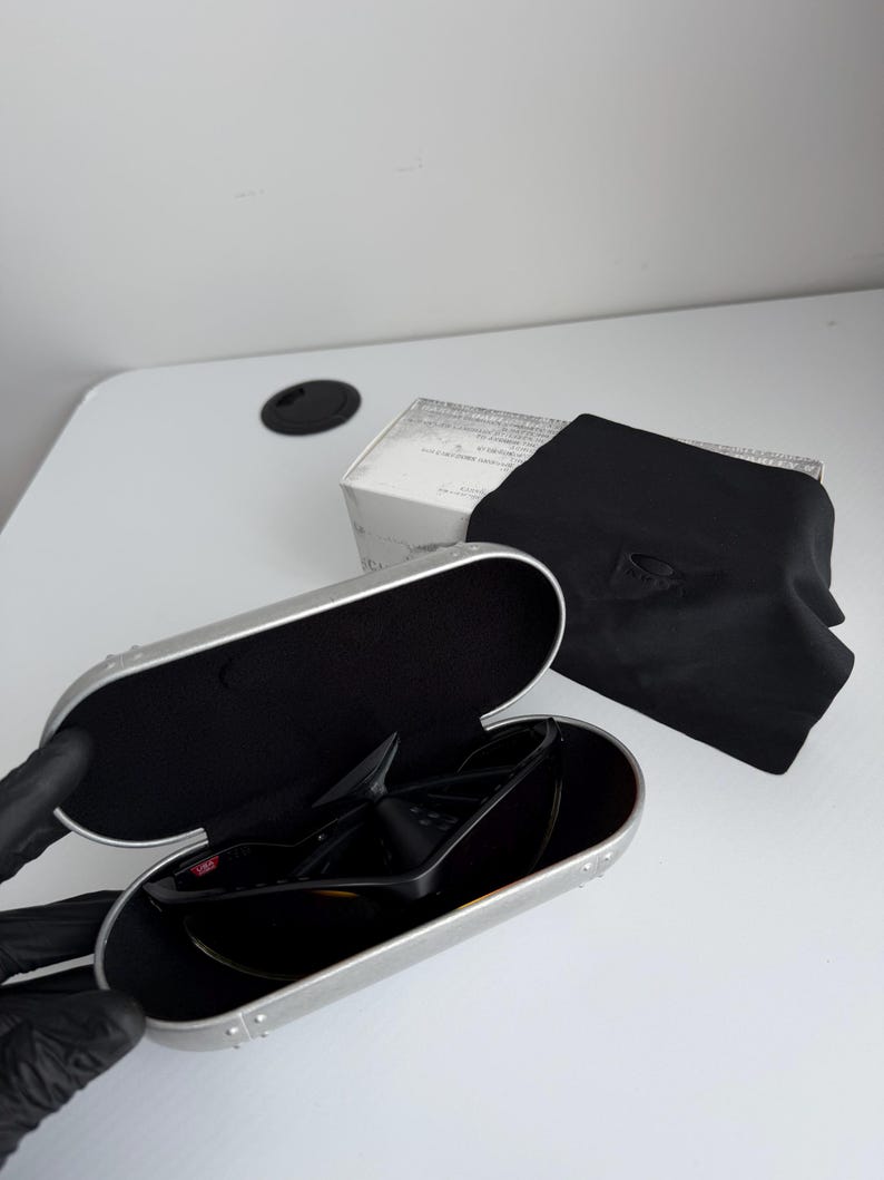 May include: Black sunglasses with a silver case, a black microfiber pouch, and a white box. The sunglasses are in an open silver case with a black interior. The black pouch has the Oakley logo.