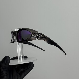 May include: Black sunglasses with purple lenses. The sunglasses have a sleek, modern design with a curved frame and arms. The lenses are a deep purple color, and the frame is a matte black. The sunglasses are displayed on a clear stand.