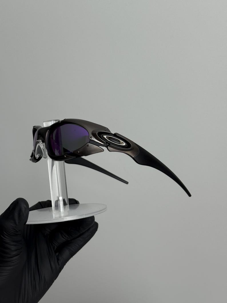 May include: Black sunglasses with purple lenses. The sunglasses have a sleek, modern design with a curved frame and arms. The lenses are a deep purple color, and the frame is a matte black. The sunglasses are displayed on a clear stand.