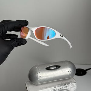 May include: White-framed sunglasses with orange and blue mirrored lenses. The sunglasses are held above a silver case and a box with the text "SCIENCE AND ART". The lenses reflect the surrounding light.