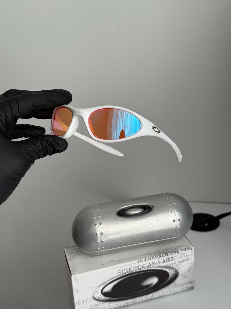 May include: White-framed sunglasses with orange and blue mirrored lenses. The sunglasses are held above a silver case and a box with the text "SCIENCE AND ART". The lenses reflect the surrounding light.