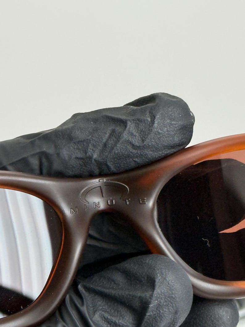 May include: Close-up of brown sunglasses with a tortoise shell pattern. The sunglasses have a curved design and the word "MINUTE" is visible on the frame. The lenses are a dark color.