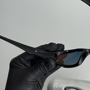 May include: Black sunglasses with a sleek, modern design. The lenses have a gradient effect, transitioning from a dark color to a lighter hue. The frames are matte black, and the arms are thin and angular.
