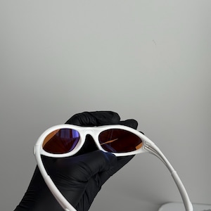 May include: White sunglasses with orange-tinted lenses. The sunglasses have a sleek, modern design with a curved frame and arms. The lenses are a warm orange color, and the frame is a bright white.