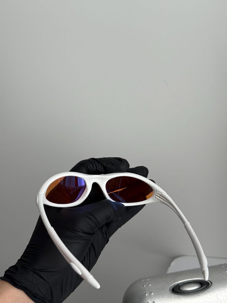 May include: White sunglasses with orange-tinted lenses. The sunglasses have a sleek, modern design with a curved frame and arms. The lenses are a warm orange color, and the frame is a bright white.