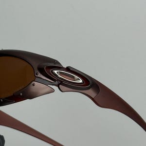 May include: Close-up of brown sunglasses with a metallic frame. The sunglasses have a curved design with a silver accent near the hinge and brown tinted lenses. The arms of the sunglasses are a darker brown.