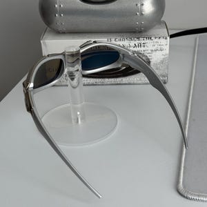 May include: Silver-framed sunglasses with blue lenses are displayed on a clear stand. A silver case with the Oakley logo is behind the sunglasses. A white box with text is behind the case.