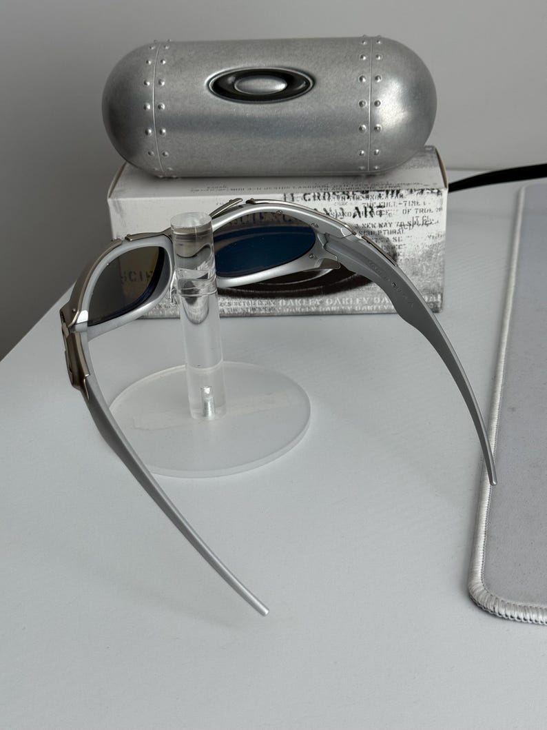 May include: Silver-framed sunglasses with blue lenses are displayed on a clear stand. A silver case with the Oakley logo is behind the sunglasses. A white box with text is behind the case.