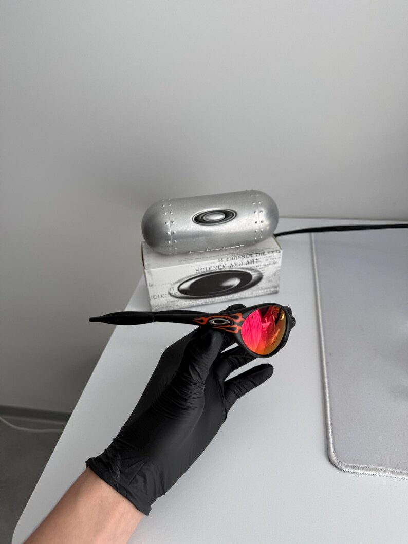 May include: Black sunglasses with red mirrored lenses and a flame design on the temples. The sunglasses are held by a gloved hand, with a silver case and a box in the background.