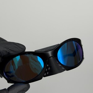 May include: Black sunglasses with blue mirrored lenses. The sunglasses have a wraparound design with a thick frame and side shields. The lenses reflect a gradient of blue and purple hues.
