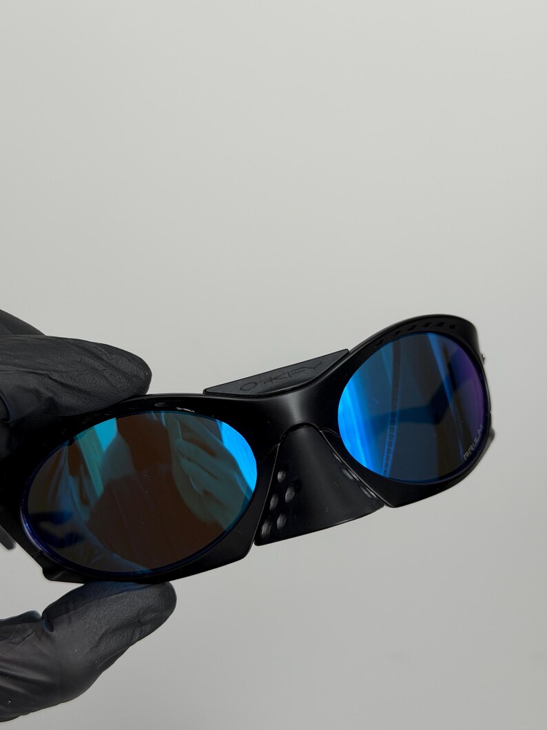 May include: Black sunglasses with blue mirrored lenses. The sunglasses have a wraparound design with a thick frame and side shields. The lenses reflect a gradient of blue and purple hues.