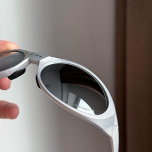 May include: Silver sunglasses with dark lenses. The sunglasses have a sleek, modern design with a curved frame and a matte finish. The lenses reflect the surrounding environment. The text "PRIZM R" is visible on the lens.
