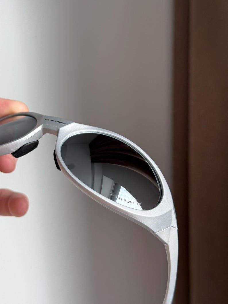 May include: Silver sunglasses with dark lenses. The sunglasses have a sleek, modern design with a curved frame and a matte finish. The lenses reflect the surrounding environment. The text "PRIZM R" is visible on the lens.