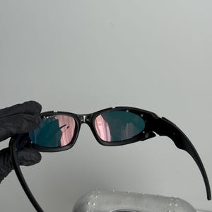 May include: Black sunglasses with a wraparound design and pink-tinted lenses. The sunglasses have a sleek, modern aesthetic. The image also shows a silver case with a black oval logo.