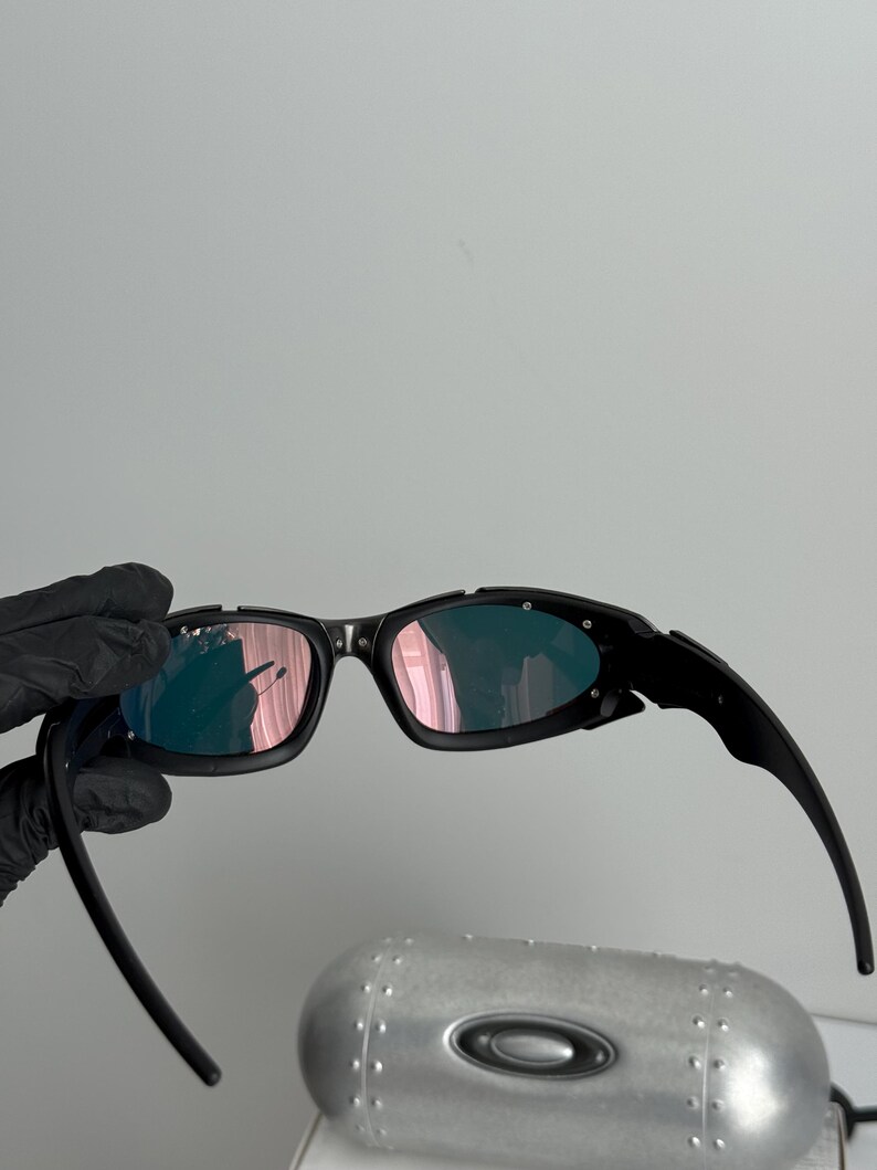 May include: Black sunglasses with a wraparound design and pink-tinted lenses. The sunglasses have a sleek, modern aesthetic. The image also shows a silver case with a black oval logo.