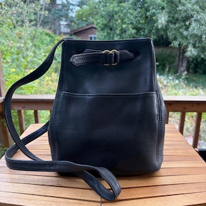 Puede incluir: Un bolso tipo cubo de cuero negro con textura granulada, que cuenta con una correa larga para el hombro y un cierre superior asegurado por un cordón de cuero fino que pasa a través de hebillas metálicas doradas. Incluye un bolsillo frontal.