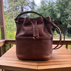 Puede incluir: Un bolso tipo cubo de cuero marrón con cierre de cordón con dos tiradores de cuero anudados, un asa superior y una correa de hombro ajustable con herrajes en tono latón. El bolso tiene una base estructurada y un acabado mate suave.