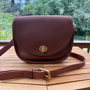 May include: A saddle-shaped brown leather crossbody bag featuring a curved front flap, a gold-toned metal turn-lock closure, and an adjustable shoulder strap with a gold-toned metal buckle. The bag has smooth, solid-colored leather construction.