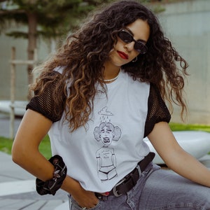 Puede incluir: Una mujer con una camiseta blanca con una ilustración en blanco y negro de una mujer con una nube y lluvia sobre su cabeza. La camiseta tiene mangas de malla negra. El texto en la camiseta dice "No siempre soy sol y arcoíris". La mujer lleva un cinturón negro y pantalones vaqueros grises.