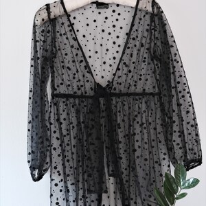 May include: Black sheer polka dot top with long sleeves and a V-neck. The top has a tie at the waist.