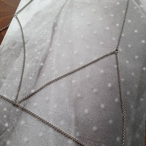 Puede incluir: Una pieza de joyería corporal de cadena plateada con múltiples cadenas conectadas que forman un patrón geométrico.