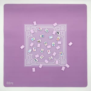May include: A square, lavender Mahjong mat with a white, ornate border design. Scattered across the mat are small, rectangular tiles and rectangular tiles with colorful illustrations. The text "the mahjong" is in the lower left corner.