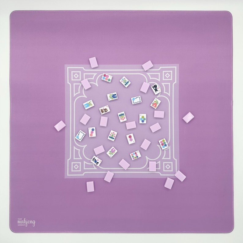 May include: A square, lavender Mahjong mat with a white, ornate border design. Scattered across the mat are small, rectangular tiles and rectangular tiles with colorful illustrations. The text "the mahjong" is in the lower left corner.