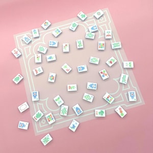 May include: A collection of white Mahjong tiles with colorful illustrations scattered on a pink surface. The tiles feature various designs, including flowers, dots, and geometric patterns. A light pink rectangular mat with a white border is visible.