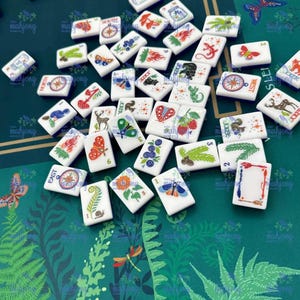 Pu&ograve; includere: Una collezione di tessere di Mahjong bianche con varie illustrazioni colorate, tra cui farfalle, fiori e bussole. Le tessere sono sparse su una superficie verde con un motivo floreale. Alcune tessere hanno numeri e la parola "JOKER".