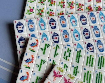 May include: A collection of white Mahjong tiles with various colorful designs. The tiles feature floral patterns, bamboo stalks, and illustrations of birds and vases. Numbers and Chinese characters are also present.
