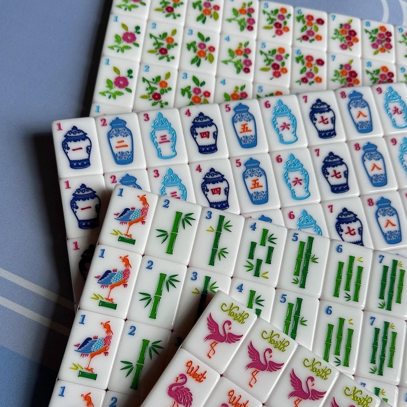 May include: A collection of white Mahjong tiles with various colorful designs. The tiles feature floral patterns, bamboo stalks, and illustrations of birds and vases. Numbers and Chinese characters are also present.