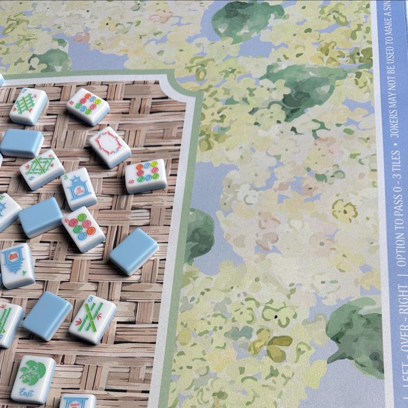 May include: A Mahjong game set with white tiles featuring various symbols and blue tiles. The game board has a floral design with text that reads "LEFT - OVER - RIGHT | OPTION TO PASS 0 - 3 TILES &bull; JOKERS MAY NOT BE USED TO MAKE A SIN".