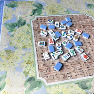 May include: A collection of Mahjong tiles with various illustrations, including boats, flowers, and lobsters, scattered on a woven mat. The tiles are white with blue accents and are arranged on a board with a floral design.
