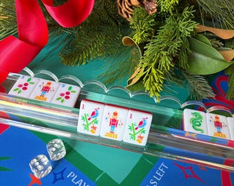 May include: A clear acrylic tile rack holding white Mahjong tiles with colorful holiday-themed graphics including nutcrackers, holly, and birds. Two transparent dice sit on a blue game mat featuring the text PLAY RIGHT and WALLS LEFT.