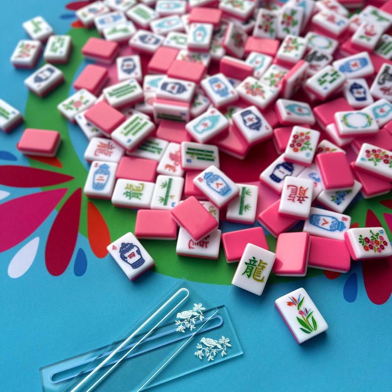 May include: A collection of pink and white Mahjong tiles scattered on a blue surface. The tiles feature various symbols and designs in green, blue, and red. Two clear acrylic tile holders with bird designs are in the foreground.