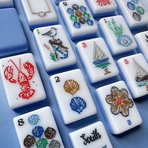May include: A collection of white mahjong tiles with light blue backs, featuring colorful engraved illustrations of nautical themes including a lobster, sailboat, seagull, anchor, seashells, and a flower, with some tiles displaying numbers or the word South.