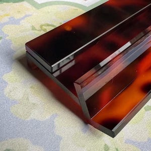 May include: A rectangular, tortoiseshell-patterned box with a hinged lid. The box has a glossy finish, with shades of brown, black, and amber. The box is set on a floral patterned surface.