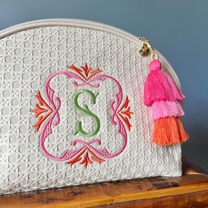 May include: Cream-colored cosmetic bag with a woven texture, featuring an embroidered pink and orange design around a green letter "S". A tri-color tassel in pink and orange hangs from the zipper.