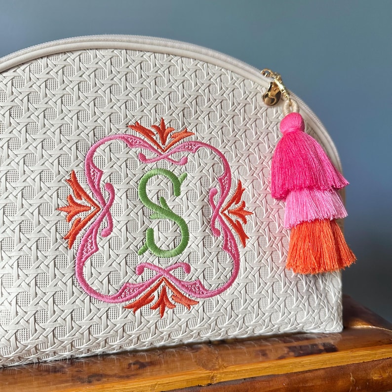 K&ouml;nnte beinhalten: Cremefarbene Kosmetiktasche mit gewebter Textur, bestickt mit einem rosa-orangenen Design um einen gr&uuml;nen Buchstaben "S". Ein dreifarbiger Quastenanh&auml;nger in Rosa und Orange h&auml;ngt am Rei&szlig;verschluss.
