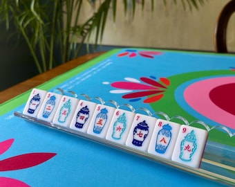 Op de afbeelding: Een set witte Mahjong-tegels met blauw-witte vaasontwerpen, genummerd van 1 tot en met 9, weergegeven in een doorzichtig plastic rek. De tegels liggen op een blauw spelbord met bloemmotieven.