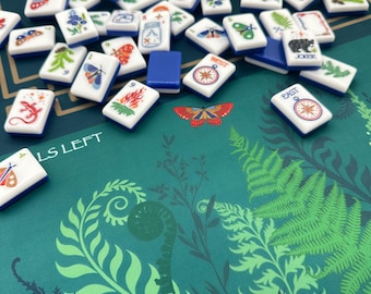 May include: A set of white rectangular mahjong tiles with blue backs, featuring colorful illustrations of insects, plants, and compasses, scattered on a dark green mat decorated with printed fern leaves and the text LS LEFT.