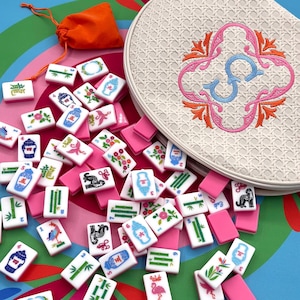 May include: A collection of white and pink Mahjong tiles scattered on a colorful surface, alongside an off-white circular case with an embroidered design and an orange drawstring bag. The tiles feature various symbols and characters.