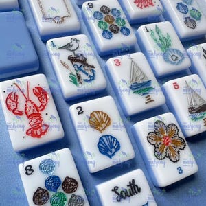 May include: A collection of white and blue Mahjong tiles with various designs, including a red lobster, a sailboat, and seashells. The tiles are arranged on a light blue surface, showcasing the detailed artwork and vibrant colors.