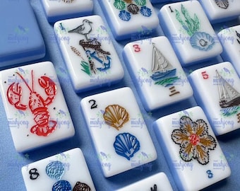 May include: A collection of white and blue Mahjong tiles with various designs, including a red lobster, a sailboat, and seashells. The tiles are arranged on a light blue surface, showcasing the detailed artwork and vibrant colors.